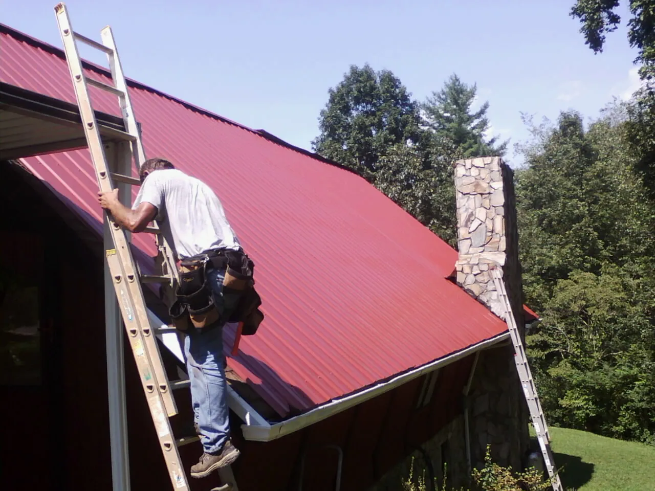 Roofer on a ladder beside a red metal roof and stone chimney — Soffit & Fascia Repair by Roofing Brothers in Pitsburg